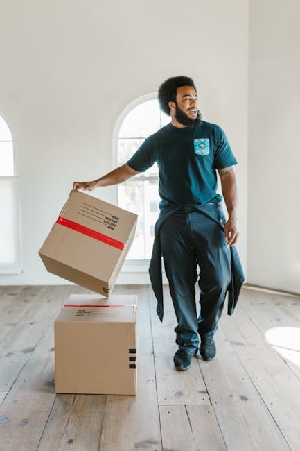 A man with dark skin, a beard, and an afro hairstyle is standing inside a property, packing items into a cardboard box labeled with a red arrow and the word 'CAUTION' on the side. He is wearing a dark