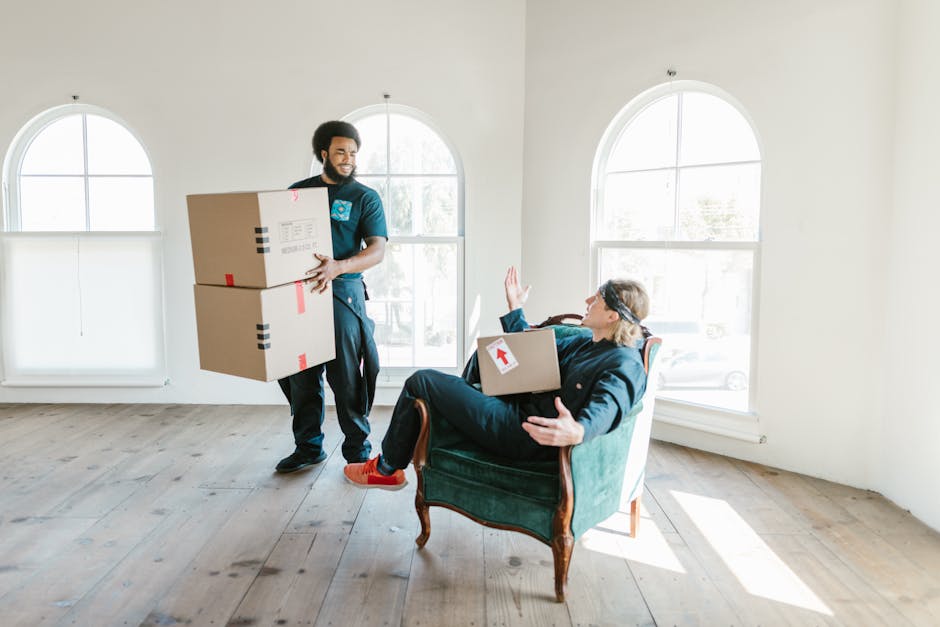 A professional removal team member in a casual dark blue uniform is inside a bright, spacious room with large arched windows that let in natural light. The team member is holding two medium-sized card