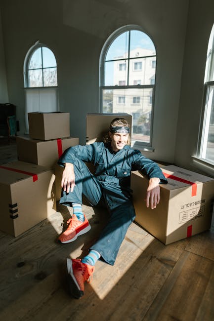 A professional removal team member in a casual dark blue uniform is inside a bright, spacious room with large arched windows that let in natural light. The team member is holding two medium-sized card