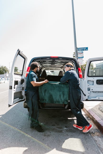 Two removal technicians loading a large piece of furniture covered with a green protective blanket into the back of a white van, which is positioned on a paved street next to a curb. The van's rear do