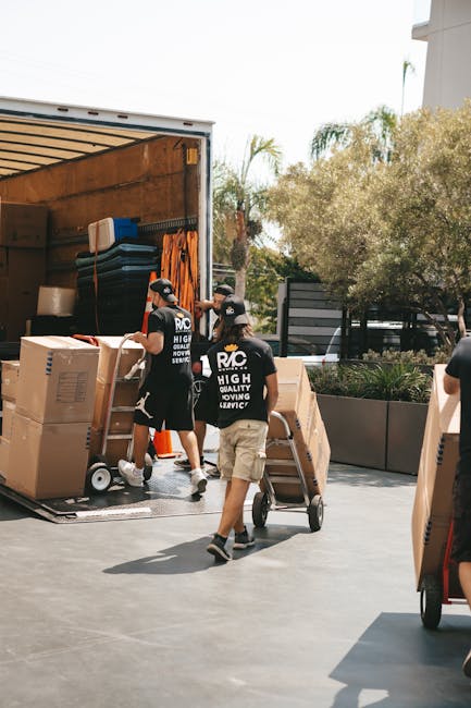 Two removal technicians loading a large piece of furniture covered with a green protective blanket into the back of a white van, which is positioned on a paved street next to a curb. The van's rear do